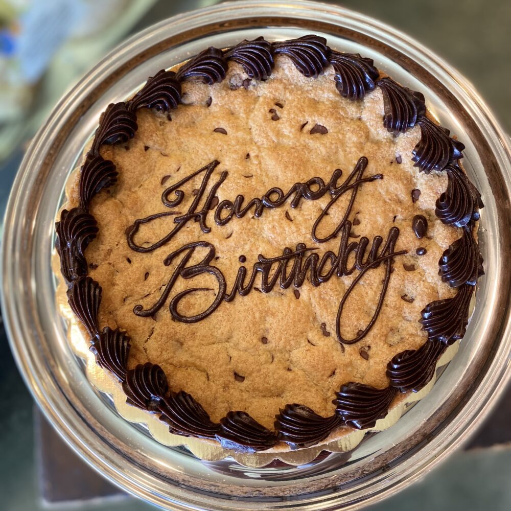 Top view of giant chocolate chip cookie with fudge frosting piped border and text reading "Happy Birthday".