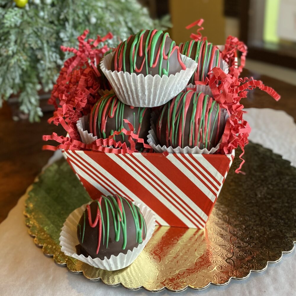 Chocolate-dipped peanut butter balls decorated with green and red chocolate drizzle in a festive gift box on a table.